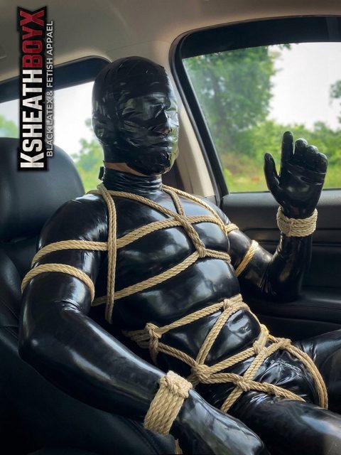 A person clad in a full black latex suit and mask sits tightly bound in rope inside a vehicle.
