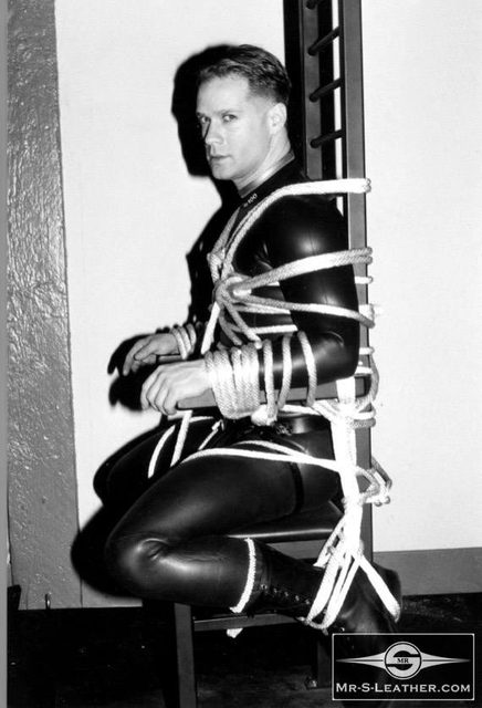 A man wearing a black rubber suit is tied securely with ropes to a chair and ladder frame in a dungeon setting.