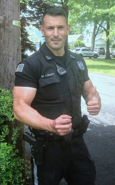 A muscular man in a black police uniform and duty belt poses outdoors with a thumbs up.