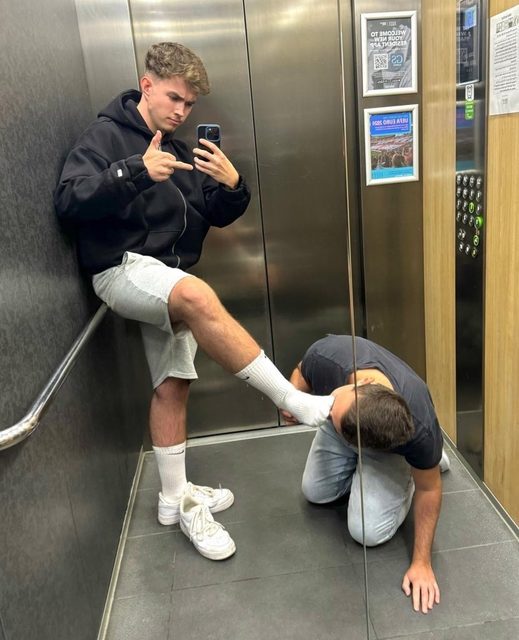 A young man takes a mirror selfie in an elevator while holding his socked foot up for another kneeling man to worship.