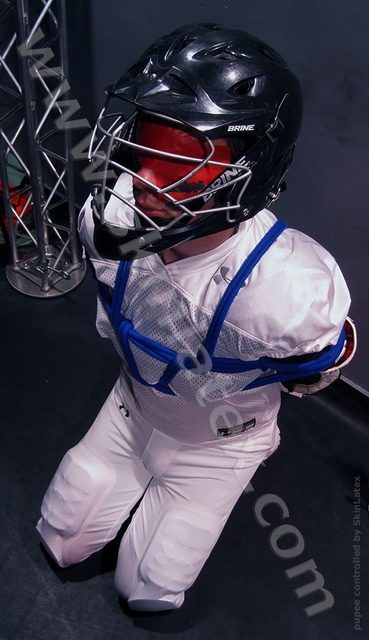 A person in a white football kit and lacrosse helmet is bound with blue rope and kneeling in a dungeon setting.