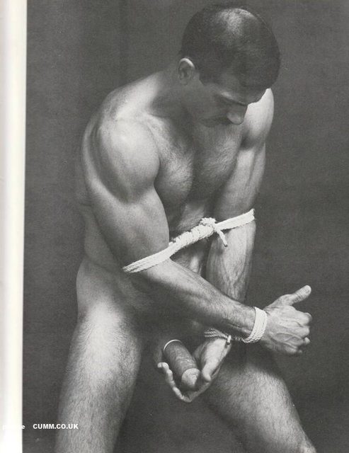 A muscular man with a hairy chest stands nude, bound by white rope around his biceps and wrists.