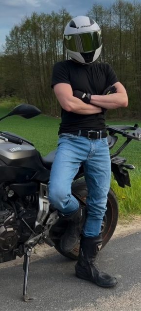 A muscular man in a motorcycle helmet, denim jeans, and leather boots poses next to his Yamaha bike outdoors.