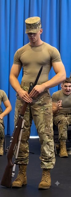 A muscular young man in a tight military t-shirt and camouflage pants poses with a rifle and bayonet.