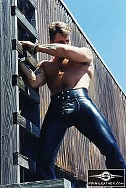 A shirtless muscular man wears tight black leather pants and wrist cuffs while posing against a wooden ladder outdoors.