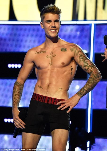 A shirtless man wearing boxer briefs and tattoos stands on a stage holding a microphone.