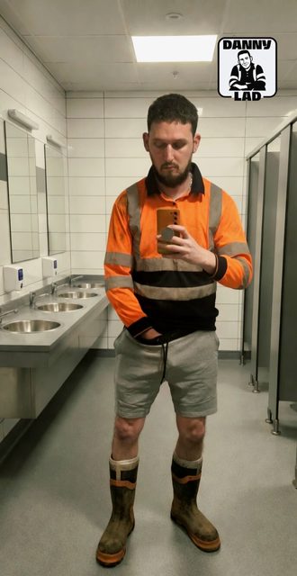 A bearded man wearing a high-visibility work shirt and rubber boots poses for a mirror selfie with his hand inside his shorts in a public restroom.