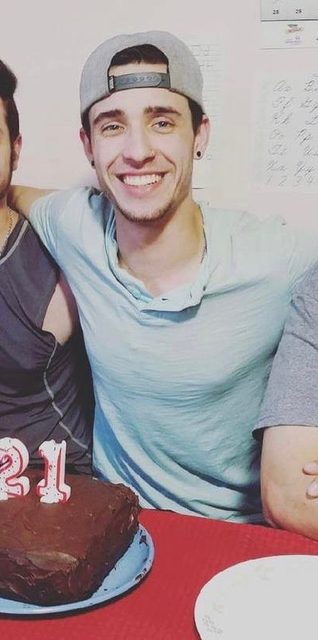 A young man with a backwards baseball cap smiles behind a 21st birthday cake.
