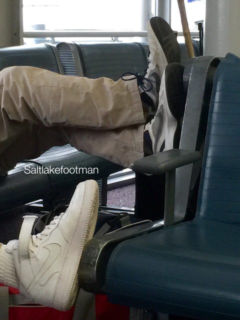 A man rests his feet on a terminal seat wearing white high-top sneakers and tube socks while another passenger rests their sneakers on a nearby armrest.