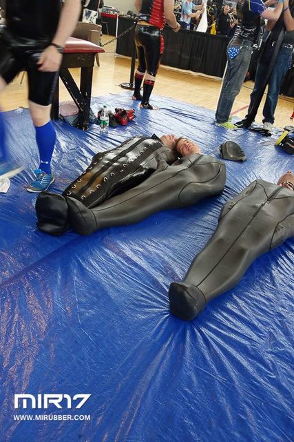 Several men are encased in heavy full-body rubber suits and sleep bags while lying on a blue tarp at a fetish event.