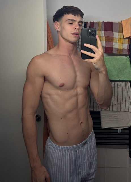 A lean, muscular young man takes a shirtless mirror selfie in a bathroom wearing striped pajama bottoms.
