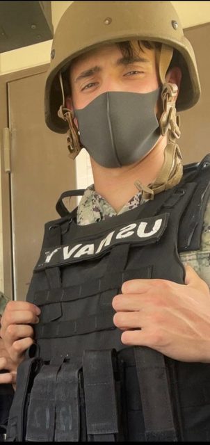 A young man wearing a US Navy tactical vest, camouflage uniform, face mask, and helmet looks at the camera.