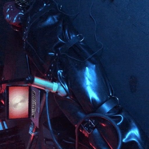 A person fully enclosed in a shiny black latex bondage sack is tightly bound with leather straps in a dimly lit dungeon setting.