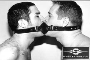 Two shirtless men are joined mouth-to-mouth wearing a shared leather gag in a black and white close-up profile view.