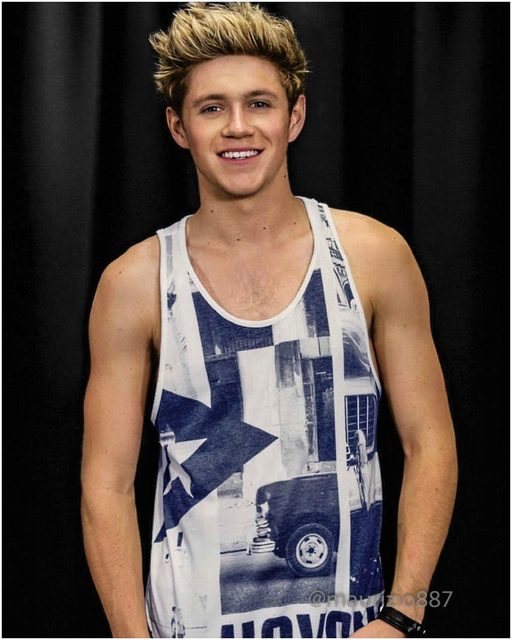 A young blonde man with spiky hair wearing a graphic tank top poses smiling against a black backdrop.