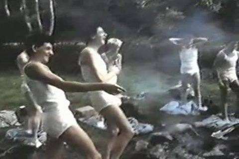 A group of slim young men in white tops and briefs dance outdoors near smoke.