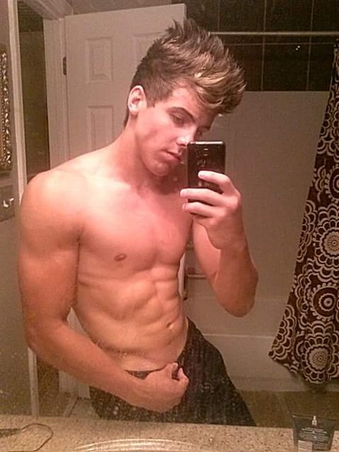 A muscular young man with a spiked hairstyle takes a shirtless mirror selfie in a bathroom, showing off his defined abs and arms.