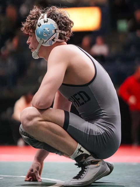 A lean young athlete wearing a grey wrestling singlet and headgear crouches on a mat in an arena.