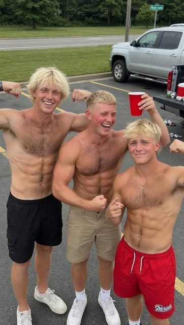 Three muscular young men with defined abs and chest hair flex their muscles while posing together outdoors in a parking lot.