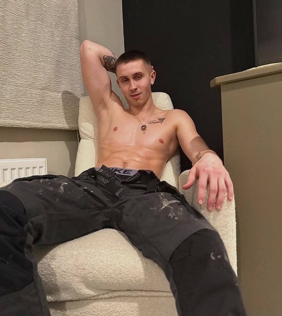 A shirtless twunk with a buzzcut and tattoos sits in an armchair with his paint-splattered work pants unbuttoned to reveal his underwear.