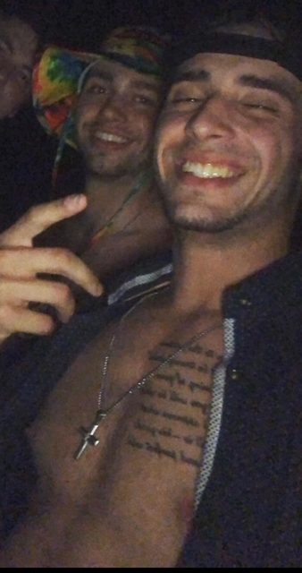 A young man with a script tattoo on his chest smiles for a selfie with friends in a dark outdoor setting.