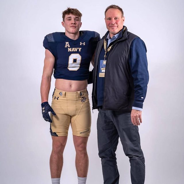 A muscular young man in a cropped Navy football uniform poses beside a mature man against a plain white background.