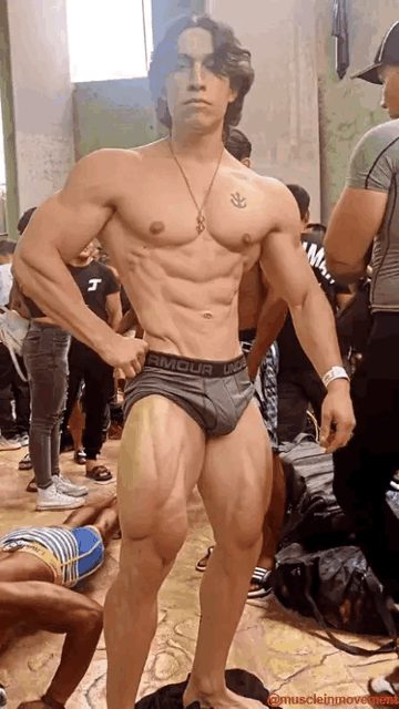 A shirtless, highly muscular young bodybuilder poses in grey Under Armour trunks while surrounded by people in a backstage or gym setting.
