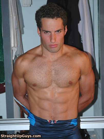 A muscular jock with a hairy chest stands in a locker room while pulling down his blue wrestling singlet.