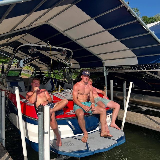 Three shirtless, muscular young men in swim trunks relax together on the back of a boat docked under a canopy.