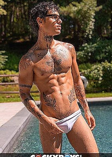 A tattooed, athletic man wearing white swim briefs poses outdoors next to a swimming pool.