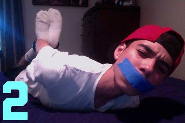 A young man wearing a white t-shirt, red cap, and white socks lies on his stomach on a bed with blue tape covering his mouth.