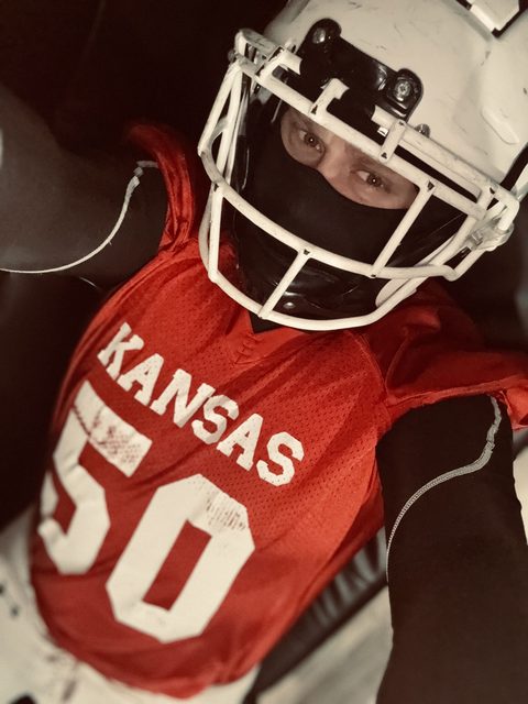 A man wears a red football jersey and a white helmet with a black balaclava covering his face.