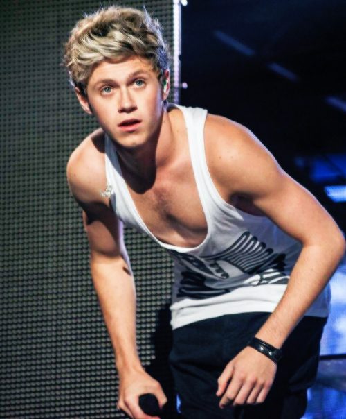 A lean, smooth young man wearing a low-cut white singlet and a black leather cuff stands on a stage.