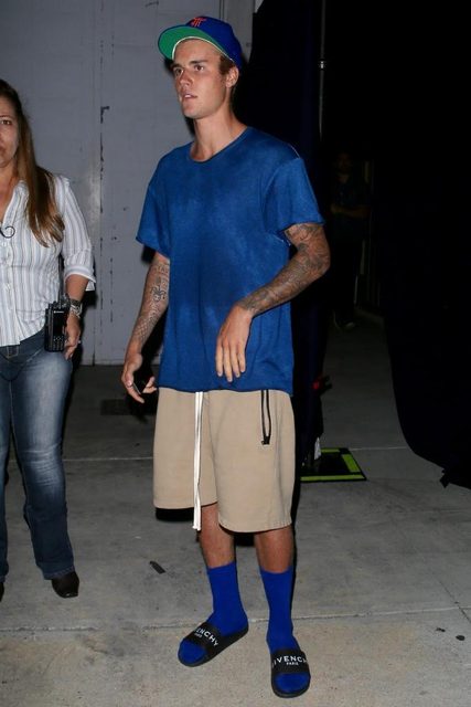 A tattooed young man stands indoors wearing a blue t-shirt, tan shorts, blue socks, and designer slides.