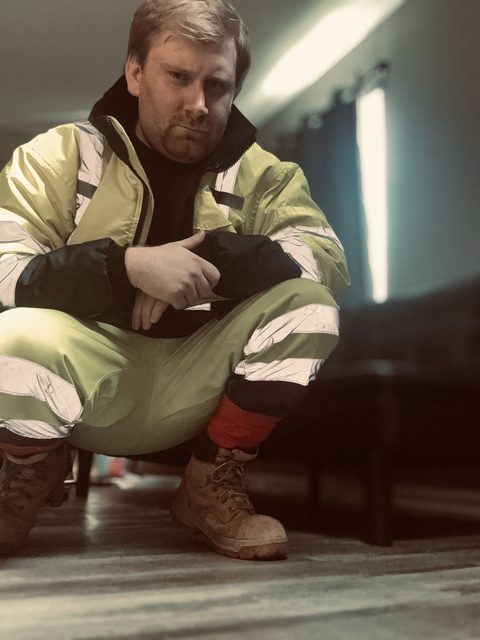 A bearded man wearing a full high-visibility construction uniform and work boots squats in an indoor setting.