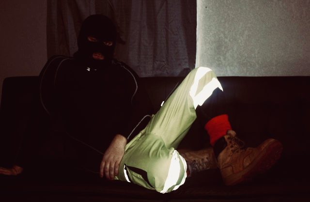 A man wearing a black balaclava and high-visibility construction trousers sits on a dark couch.