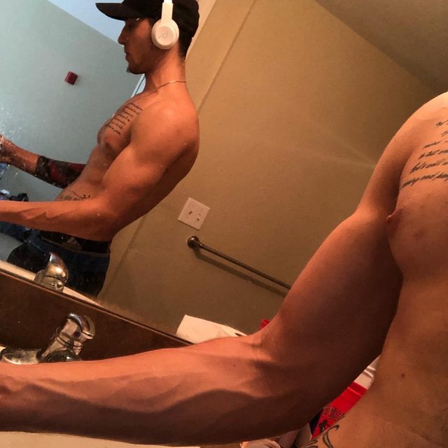 A shirtless, tattooed man takes a mirror selfie in a bathroom, showcasing his vascular arms and muscular build while wearing a baseball cap and headphones.