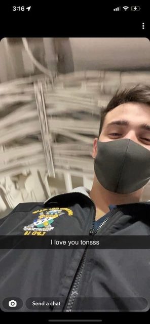 A young man in a naval utility jacket and face mask stands in a ship's interior with visible overhead piping.
