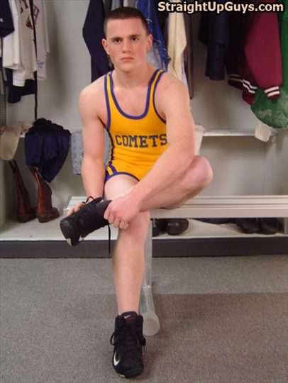 A young man with a buzzcut wearing a yellow and purple wrestling singlet sits on a bench in a locker room while putting on black sneakers.