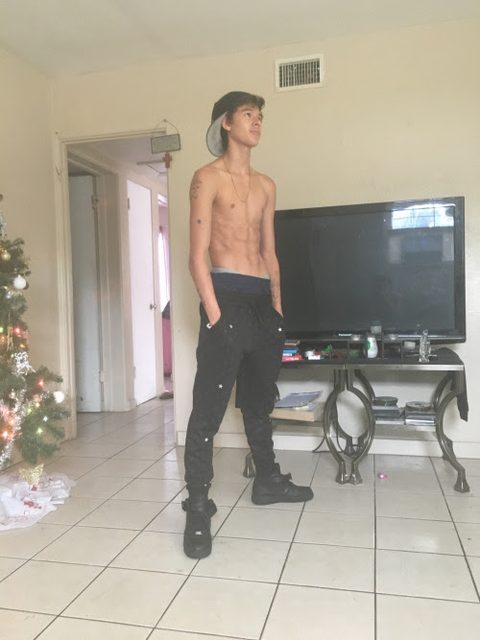 A lean shirtless twink poses in a living room wearing sagging black joggers and a backward cap.