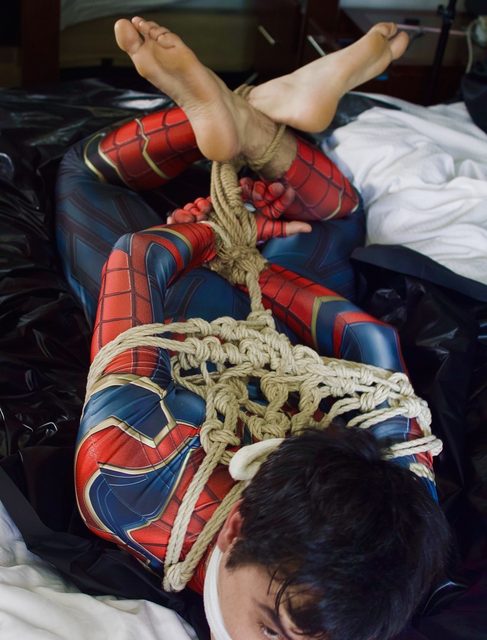 A man in a Spidey-themed spandex suit is bound in complex rope bondage and gagged while lying on a bed.