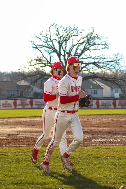 Two athletic young men wearing matching red and white baseball uniforms jog across a sunlit baseball field.