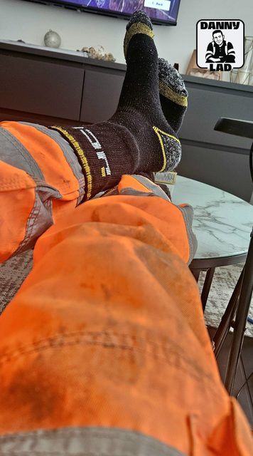 A point-of-view shot features a man's legs in stained orange high-visibility trousers and black socks resting on a table.