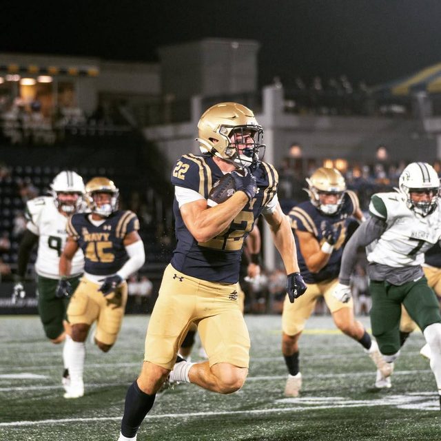 A muscular football player in a navy and gold uniform runs with the ball on the field during a night game.