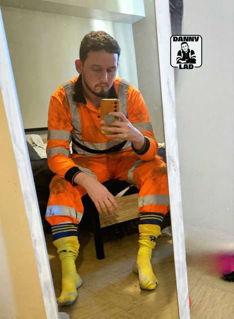 A bearded man wearing a dirty orange hi-vis boiler suit and long yellow football socks takes a mirror selfie in a bedroom.