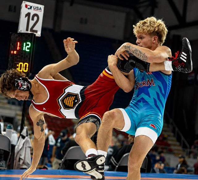 Two young wrestlers in tight athletic singlets engage in a high-impact grappling match on a competition mat in an arena.