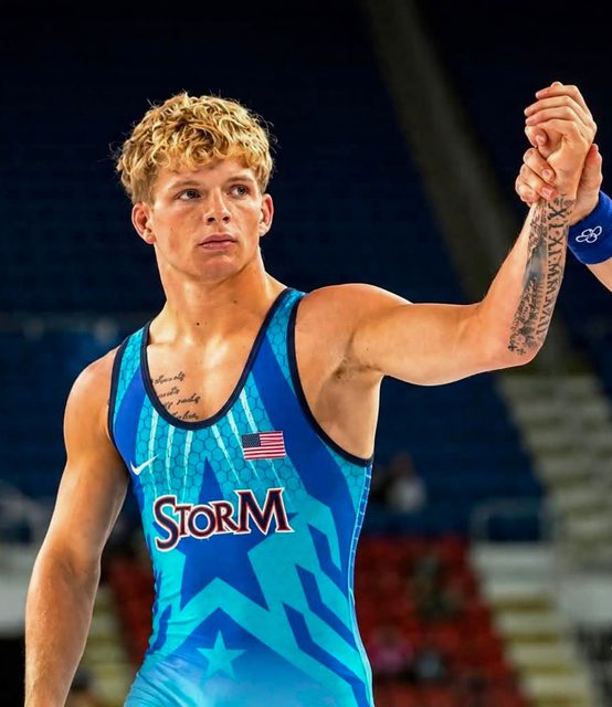 A young athletic man with blond curly hair and tattoos wears a blue patterned wrestling singlet as his arm is raised in a wrestling arena.