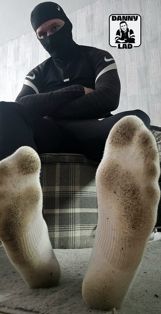 A man wearing black athletic gear and a balaclava sits with his legs open, showing the soles of his dirty white socks to the camera.