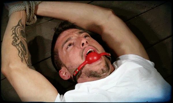 A muscular man with a forearm tattoo lies on a wooden floor with his wrists bound by rope and a red ball gag in his mouth.