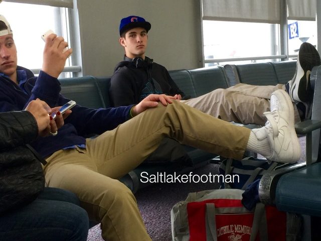 Several young men in casual attire, including khakis and hoodies, wait in an airport terminal, highlighting a focus on their white sneakers and tube socks.